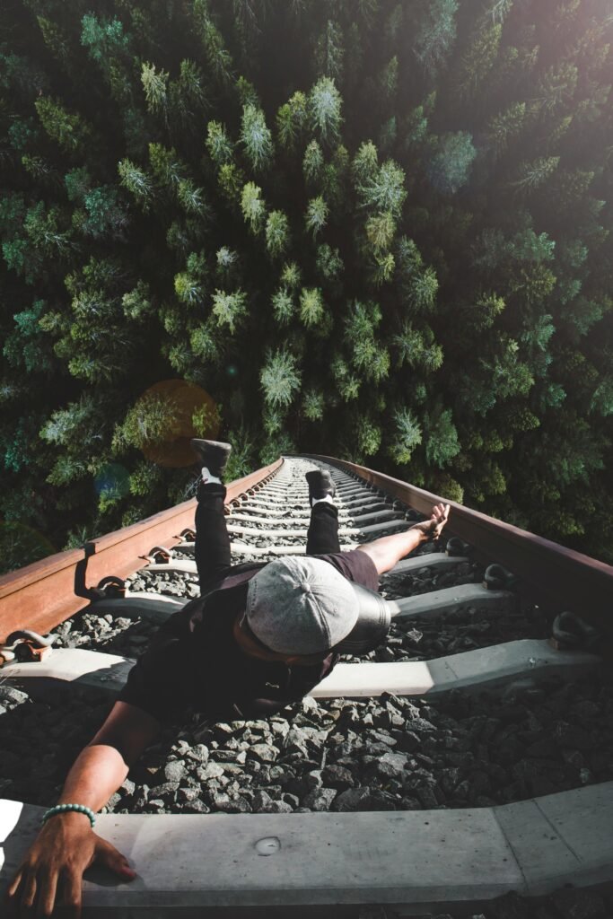 A man poses daringly over train tracks, as if he is falling from great hights into the forest below, at the end of the tracks.